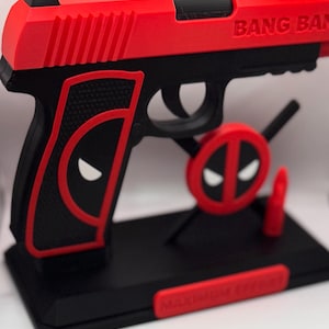 May include: A red and black 3D-printed handgun with the text "BANG BANG" on the side. The design incorporates the Deadpool logo and a stand. The base has the text "MAXIMUM EFFORT".