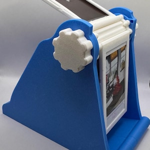 May include: A blue and white photo display stand with a built-in frame. The stand has a blue base and a white frame that holds a photo. The stand has a gear-shaped knob on the side. The top of the stand has the word "Instax".