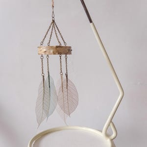 May include: A decorative mobile featuring delicate, translucent leaf shapes suspended from a wooden ring by chains. The mobile hangs from a cream-colored metal stand with a circular base, creating a minimalist, natural aesthetic.