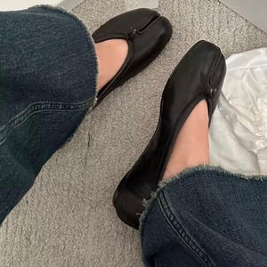 May include: Black leather split-toe ballet flats. The shoes have a unique design with a separate section for the big toe. The flats are paired with blue denim jeans. The shoes are a stylish and modern footwear choice.