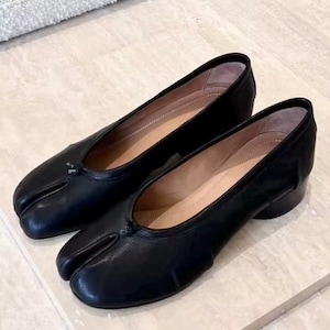 May include: A pair of black leather split-toe flats with a low heel. The shoes have a distinctive design with a separate section for the big toe. The interior is a light tan colour, and they are displayed on a light-coloured surface.