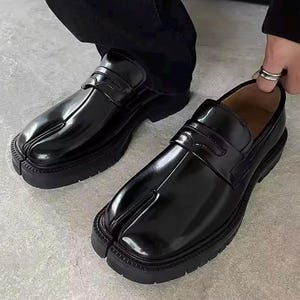 May include: Pair of black leather split-toe loafers with a chunky sole. The shoes have a glossy finish and a strap detail across the top. The design features a unique split-toe construction, offering a distinctive look.