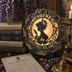 Könnte beinhalten: Eine kreisförmige, dunkel gehaltene Zierplatte mit einem verzierten Rahmen im Filigranstil und einer hinterleuchteten Silhouette einer Person mit Federkiel. Der Text "DEAREST GENTLE READER" ist am Rand eingeprägt. Die Platte ist beleuchtet.