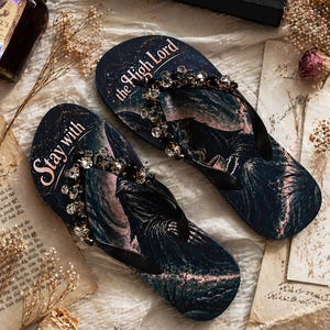 Puede incluir: Un par de chanclas azul oscuro con una banda de cristal y un diseño de paisaje. Las palabras "Stay with" y "the High Lord" están impresas en las chanclas. También se ve un libro titulado "A Court of Thorns and Roses".