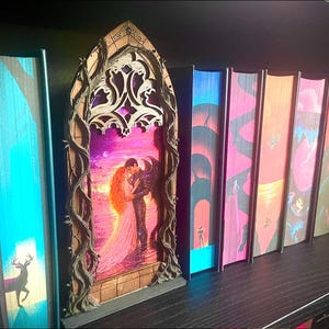 Op de afbeelding: Een verzameling fantasyboeken met kleurrijke ruggen en een decoratief houten element. Het houten element toont een romantische sc&egrave;ne van een omhelzend paar, omlijst door een boog en wijnranken. De boekruggen tonen diverse illustraties.