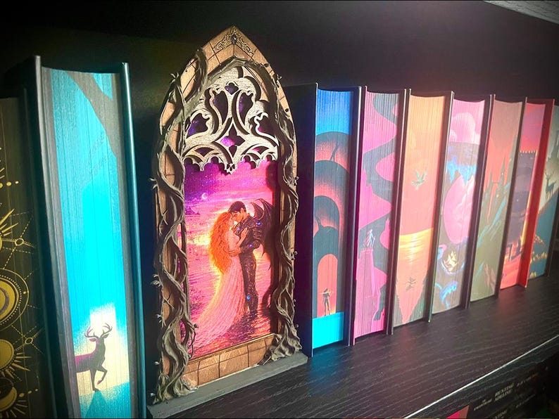 Op de afbeelding: Een verzameling fantasyboeken met kleurrijke ruggen en een decoratief houten element. Het houten element toont een romantische sc&egrave;ne van een omhelzend paar, omlijst door een boog en wijnranken. De boekruggen tonen diverse illustraties.