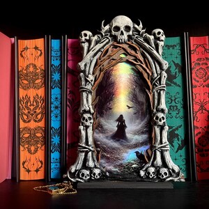 May include: A gothic-themed decorative piece featuring a skull-adorned frame with a mystical forest scene. The frame is flanked by colorful books with intricate designs. A gold-colored pendant rests in front of the frame, adding a touch of elegance to the dark aesthetic.