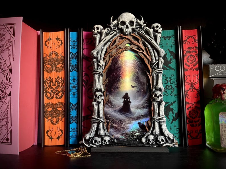 May include: A gothic-themed decorative piece featuring a skull-adorned frame with a mystical forest scene. The frame is flanked by colorful books with intricate designs. A gold-colored pendant rests in front of the frame, adding a touch of elegance to the dark aesthetic.