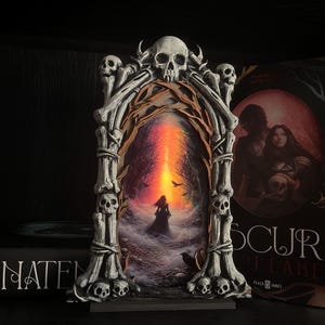 May include: A gothic-style decorative frame with a skull and bone design, surrounding a painting of a figure in a dark landscape with a fiery sky. A book with the title "OSCURA" is visible.