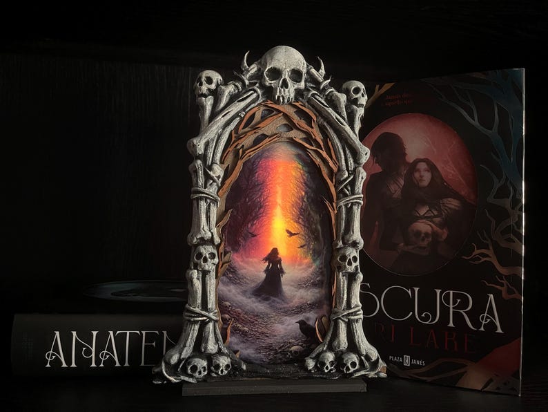 May include: A gothic-style decorative frame with a skull and bone design, surrounding a painting of a figure in a dark landscape with a fiery sky. A book with the title "OSCURA" is visible.