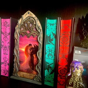 Op de afbeelding: Een verzameling decoratieve boeken met kleurrijke ruggen, waaronder oranje, blauw en roze. Een gesneden houten frame met een romantische zonsondergangsc&egrave;ne staat centraal. Een gouden masker en een decoratieve fles zijn ook zichtbaar.