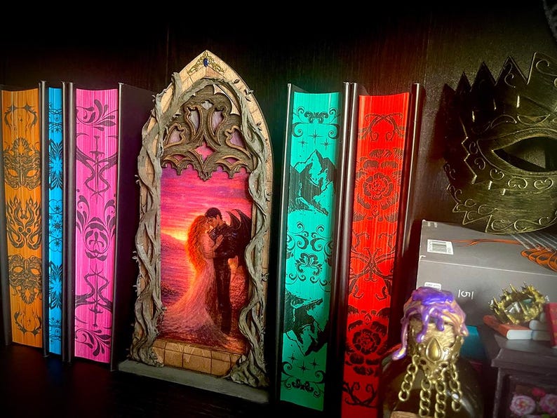 Op de afbeelding: Een verzameling decoratieve boeken met kleurrijke ruggen, waaronder oranje, blauw en roze. Een gesneden houten frame met een romantische zonsondergangsc&egrave;ne staat centraal. Een gouden masker en een decoratieve fles zijn ook zichtbaar.