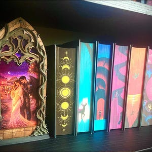 Op de afbeelding: Een decoratief, boogvormig houten frame met een verlicht beeld van een stel in een romantische sc&egrave;ne. Daarnaast een rij kleurrijke boeken met unieke omslagontwerpen, op een plank.