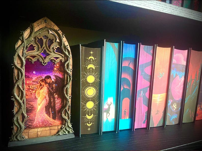 Op de afbeelding: Een decoratief, boogvormig houten frame met een verlicht beeld van een stel in een romantische sc&egrave;ne. Daarnaast een rij kleurrijke boeken met unieke omslagontwerpen, op een plank.