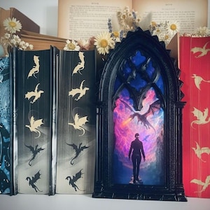 Puede incluir: Una obra de arte enmarcada en negro de estilo gótico con una escena de dragones, flanqueada por libros decorativos. Los libros tienen motivos de dragones y pájaros en oro y blanco sobre fondos oscuros. Flores secas y páginas de libros antiguos visibles.