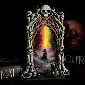 May include: A gothic-style decorative frame with a skull and bone design. The frame surrounds a painting of a woman in a dark forest with a rainbow-colored light. A book with the word "ANATEM" is visible.