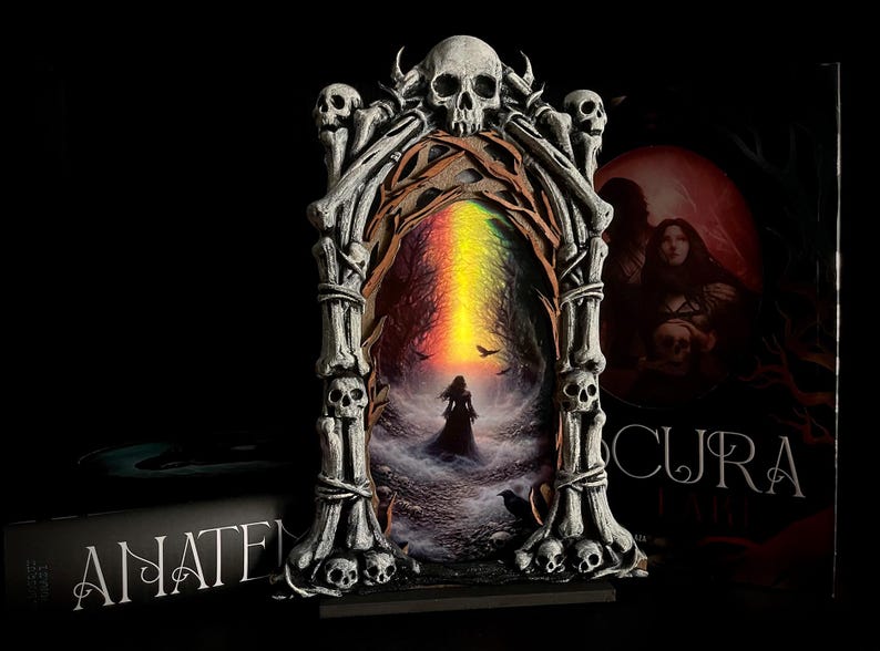 May include: A gothic-style decorative frame with a skull and bone design. The frame surrounds a painting of a woman in a dark forest with a rainbow-colored light. A book with the word "ANATEM" is visible.