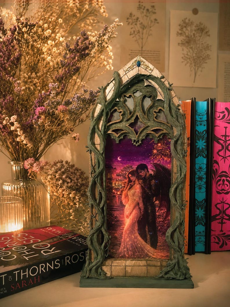 Op de afbeelding: Een decoratief gotisch frame met een romantische sc&egrave;ne van een stel. Het frame is groen met een wijnstokachtig ontwerp. Een boek met de titel "A Court of Thorns and Roses" is zichtbaar. Droogbloemen en een kaars staan ook in de afbeelding.