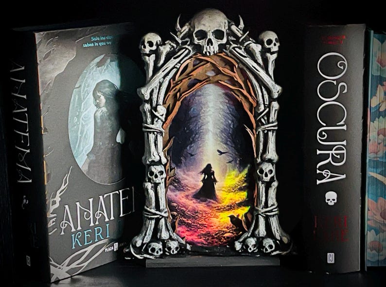 May include: A decorative, gothic-style archway with a skull at the top and smaller skulls along the sides. The archway frames a colorful painting of a silhouetted figure in a dark landscape. Flanking the arch are two books with titles in Spanish.