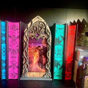 Op de afbeelding: Een verzameling fantasyboeken met decoratieve randen in oranje, blauw, roze, teal en rood. Een houten kunstwerk met een omhelzend stel staat centraal. Een gouden masker en een decoratieve fles zijn ook aanwezig.