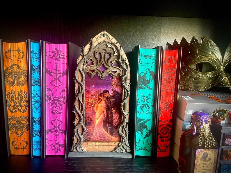 Op de afbeelding: Een verzameling fantasyboeken met decoratieve randen in oranje, blauw, roze, teal en rood. Een houten kunstwerk met een omhelzend stel staat centraal. Een gouden masker en een decoratieve fles zijn ook aanwezig.
