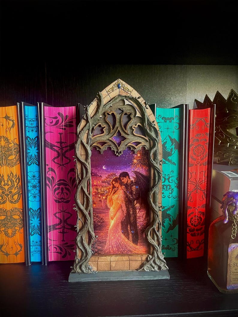 Op de afbeelding: Een decoratief frame in gotische stijl met een romantische afbeelding van een stel, tegen een achtergrond van kleurrijke boeken. Het frame is ingewikkeld ontworpen met een donkerbruine afwerking en de boeken hebben levendige covers in oranje, blauw, roze, groen en rood.