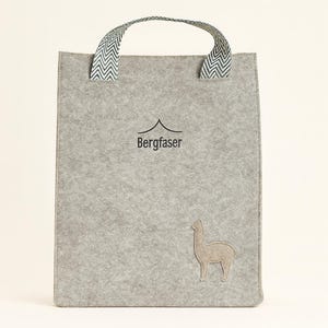 Könnte beinhalten: Eine graue Filz-Tragetasche mit einem Griff mit Zickzackmuster. Die Tasche trägt den Schriftzug "Bergfaser" und ein gesticktes Lama-Design. Die Tasche ist rechteckig und hat ein schlichtes, minimalistisches Design.