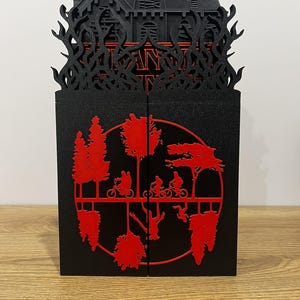 Puede incluir: Caja decorativa negra y roja cortada con láser con una silueta de casa y la palabra "Stranger Things". La caja tiene un diseño rojo y negro con una escena de personas en bicicleta y árboles invertidos.
