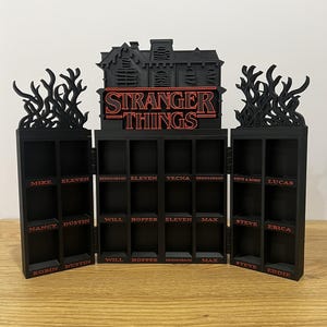 Puede incluir: Estantería negra de Stranger Things con múltiples compartimentos. La parte superior presenta una silueta de casa y el logotipo del programa en rojo. Los nombres de los personajes como Mike, Eleven y Dustin están etiquetados.