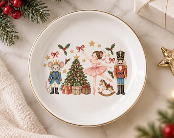 Può includere: Un piatto rotondo in ceramica bianca con bordo dorato, caratterizzato da un'illustrazione colorata di un albero di Natale, una ballerina in tutù rosa, uno schiaccianoci, un topo in uniforme, un cavallo a dondolo e regali circondati da agrifoglio.