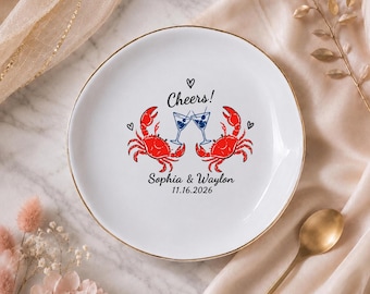 Può includere: Un piatto in ceramica bianca con bordo dorato raffigurante due granchi rossi che tengono in mano bicchieri da martini blu. Il testo recita Cheers! Sophia & Waylon 16.11.2026, accompagnato da piccole icone a forma di cuore nero.