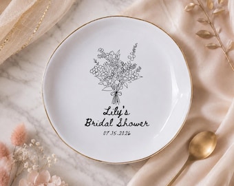 Può includere: Piatto in ceramica bianca con bordo dorato, con un design a bouquet floreale nero e la scritta "Lily's Bridal Shower 07.15.2026". Il piatto è un oggetto decorativo, adatto a un addio al nubilato.