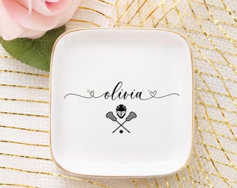 Può includere: Piatto quadrato in ceramica bianca con bordo dorato. Il piatto presenta il nome "Olivia" in corsivo, con un casco da lacrosse, bastoni incrociati e una grafica a palla. Una rosa rosa è sullo sfondo.
