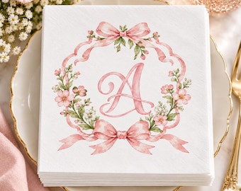 Può includere: Una pila di tovaglioli di carta quadrati bianchi con una lettera A rosa ad acquerello al centro. La lettera è circondata da una ghirlanda circolare di fiori rosa, foglie verdi e due grandi nastri rosa legati in alto e in basso.