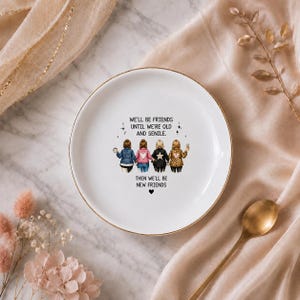 Puede incluir: Un plato decorativo redondo de cer&aacute;mica blanca con un borde dorado. El centro presenta una ilustraci&oacute;n de cuatro amigos de pie uno al lado del otro con el texto: WE'LL BE FRIENDS UNTIL WE'RE OLD AND SENILE. THEN WE'LL BE NEW FRIENDS.