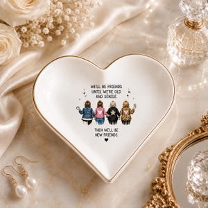 Puede incluir: Un plato de cer&aacute;mica blanco en forma de coraz&oacute;n con un borde dorado. El centro presenta una ilustraci&oacute;n de cuatro amigos vistos desde atr&aacute;s y el texto: WE'LL BE FRIENDS UNTIL WE'RE OLD AND SENILE. THEN WE'LL BE NEW FRIENDS con un peque&ntilde;o coraz&oacute;n negro.
