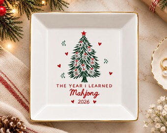 Può includere: Un piattino quadrato in ceramica bianca con bordo dorato. Al centro, un albero di Natale verde decorato con tessere del Mahjong, cuori rossi e stelline. Sotto l'albero, il testo recita THE YEAR I LEARNED Mahjong 2026.