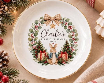 Può includere: Un piatto in ceramica bianca con bordo dorato decorato con una ghirlanda natalizia, un coniglio marrone, regali incartati e la scritta "Charlie's FIRST CHRISTMAS 2026" al centro, sormontato da un fiocco a quadretti beige e una stella dorata.