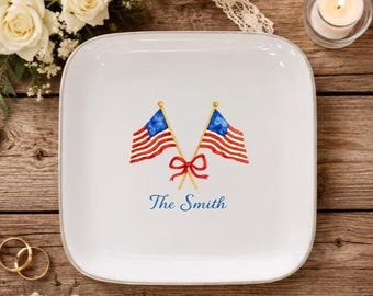 Può includere: Piatto quadrato bianco con angoli arrotondati, con due bandiere americane a strisce rosse e bianche e campi di stelle blu. Un fiocco rosso lega gli alberi delle bandiere, con il nome "The Smith" in blu sotto.