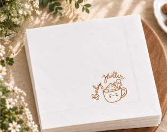 Può includere: Una pila di tovaglioli di carta quadrati bianchi con una stampa marrone raffigurante una tazza con un viso sorridente, panna montata e un omino di pan di zenzero, accompagnata dal testo corsivo marrone Baby Miller in basso a destra.