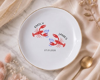 Può includere: Piatto in ceramica bianca con bordo dorato, con illustrazioni di aragoste rosse e bicchieri da cocktail blu. I nomi "EMILY" e "JAMES" sono stampati sopra le aragoste, con la data "07.01.2026" sotto. Un oggetto decorativo.