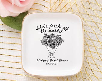 Può includere: Un vassoio quadrato in ceramica bianca con bordo dorato. Il piatto presenta il testo "She's fresh off the market" sopra un'illustrazione di bouquet. Sotto, il testo "Madison's Bridal Shower 07.15.2026".