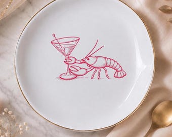 Può includere: Un piatto in ceramica bianca con bordo dorato che presenta un'illustrazione a linee rosse di un'aragosta che tiene in mano un bicchiere da martini con un'oliva. Il disegno è centrato sul piatto, mostrando un crostaceo e un bicchiere.