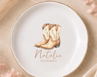 Può includere: Un piattino rotondo in ceramica bianca con bordo color oro. Il centro presenta un'illustrazione di due stivali da cowboy color marrone chiaro con dettagli floreali. Sotto gli stivali, il testo riporta Natalie in corsivo e BRIDESMAID in stampatello.