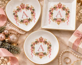 Può includere: Tre piattini in ceramica bianca con bordi dorati, che includono una forma a cuore, una quadrata e una rotonda. Ogni piattino presenta una lettera A centrale circondata da una ghirlanda decorativa di nastri rosa, una corona e del fogliame.