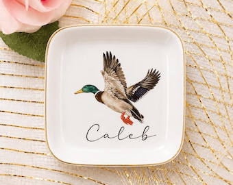 Può includere: Un piattino quadrato in ceramica bianca con bordo dorato. Il centro presenta un'illustrazione dettagliata di un germano reale in volo con piume verdi, marroni e bianche. Sotto l'anatra, il nome Caleb è scritto in corsivo nero.