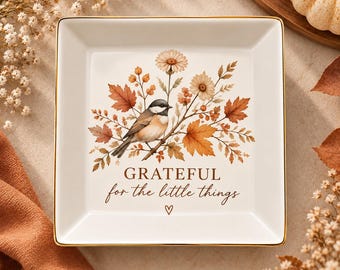 Puede incluir: Un plato cuadrado de cerámica blanca con borde dorado. El centro muestra un carbonero cabecinegro posado en una rama rodeado de hojas de otoño, bayas y flores, junto con el texto "GRATEFUL for the little things" y un pequeño corazón.