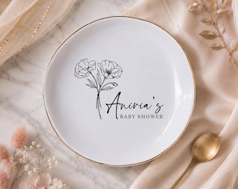 Può includere: Piatto in ceramica bianca con bordo dorato, con un design floreale nero e la scritta "Anivia's BABY SHOWER". Il piatto è appoggiato su un tessuto color crema con un cucchiaio dorato e accenti floreali.
