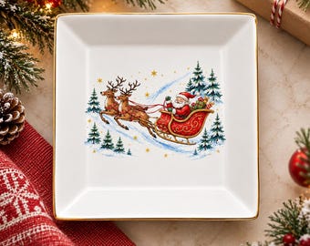 Può includere: Un piattino quadrato in ceramica bianca con bordo dorato. Il centro presenta un'illustrazione colorata di Babbo Natale su una slitta rossa trainata da due renne marroni attraverso un bosco innevato con pini verdi e stelle dorate.