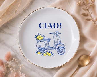 Può includere: Un piatto rotondo in ceramica bianca con bordo dorato. Il centro presenta un'illustrazione a linee blu di uno scooter con un cestino di limoni gialli, accompagnata dal testo blu "CIAO!" stampato in un carattere grassetto senza grazie.